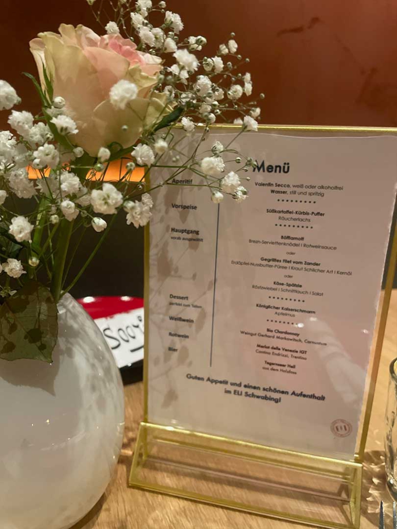Detailaufnahme einer Menükarte und Blumendekoration auf einem festlich gedeckten Tisch im Restaurant ELI Schwabing.
