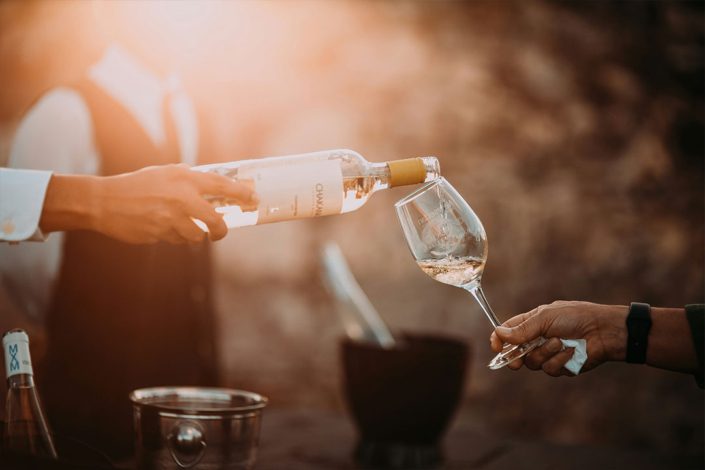 Weißwein wird aus einer Flasche in ein Weinglas eingeschenkt, warme Abendstimmung im Hintergrund.